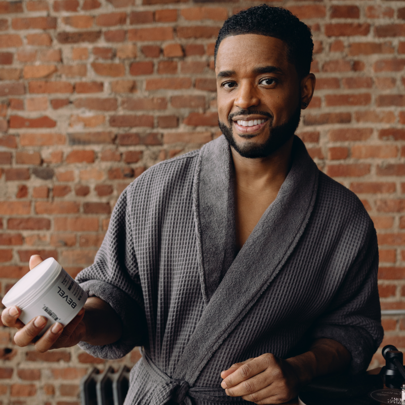 Larenz Tate in a gray robe holding a Bevel Body Cream against a brick wall