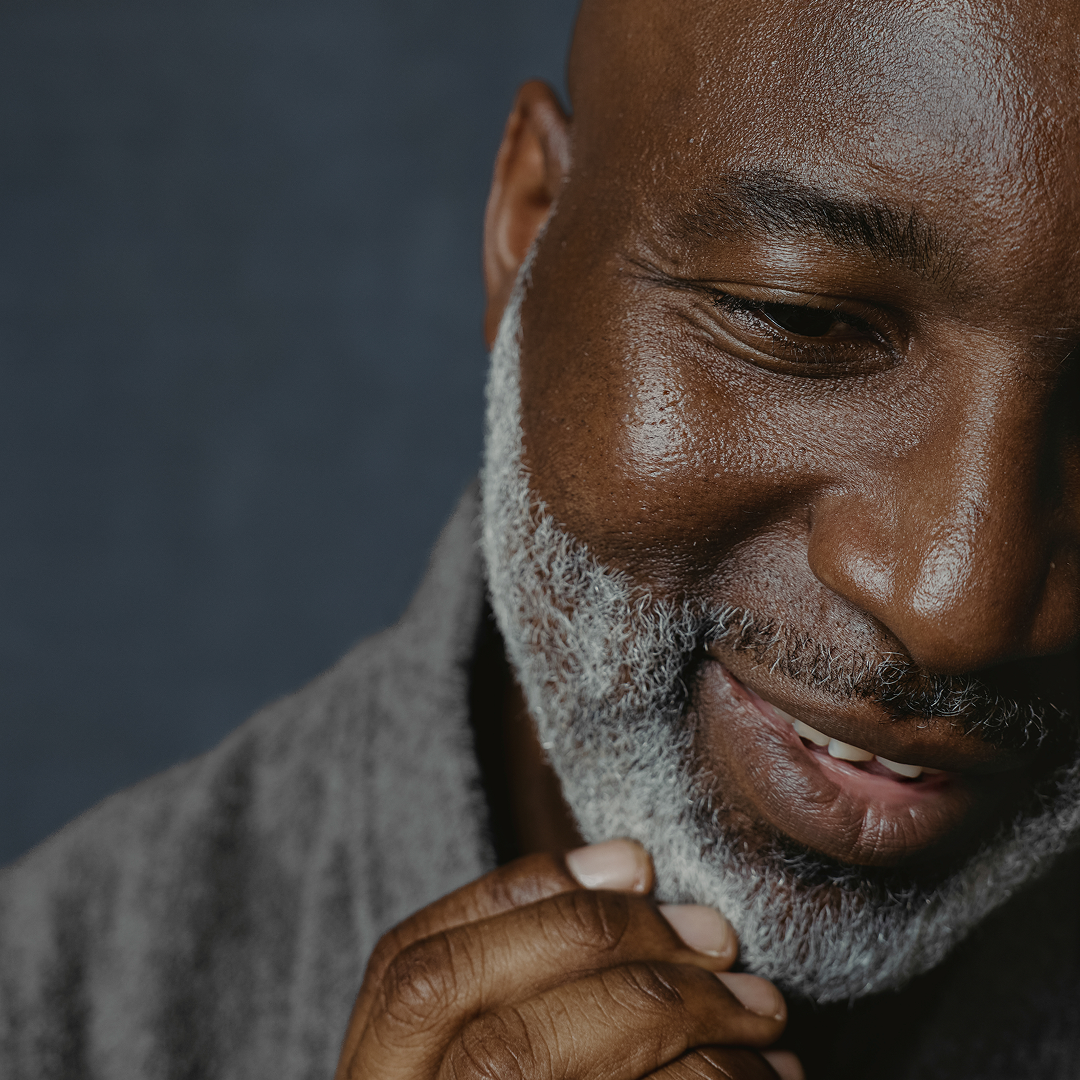Close-up of a man with a gray beard and gray sweater against a dark background