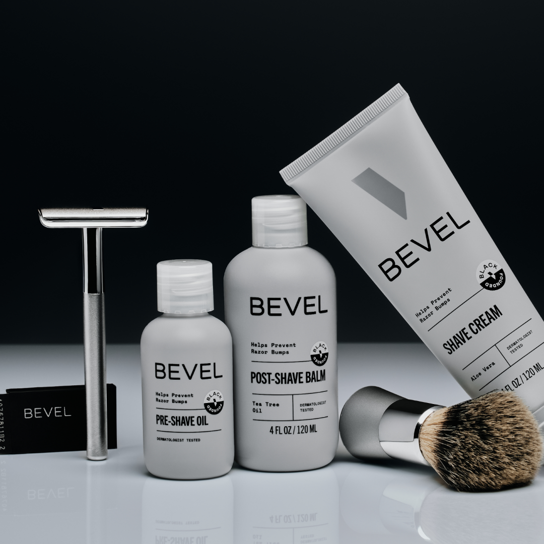 Bevel shaving products including a razor, oil, balm, and cream on a reflective surface with a black background.