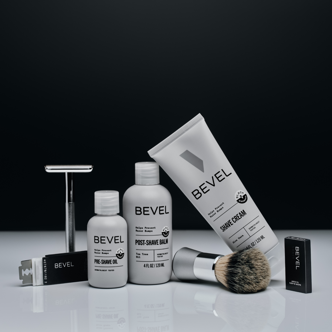 Bevel grooming products including bottles and a brush on a reflective surface with a black background